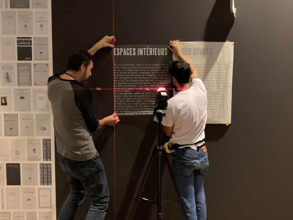 Photographie de deux personnes qui installent au mur un cartel d'exposition à l'aide d'un niveau laser