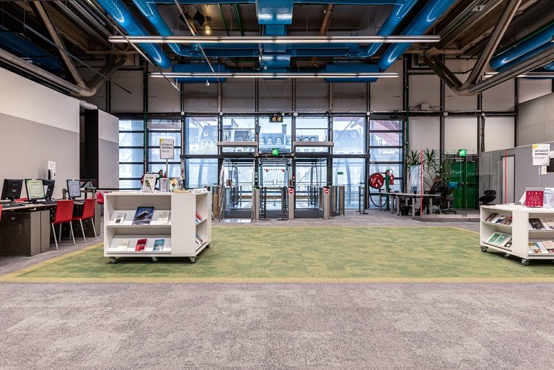 Entrée publique du niveau 2 de la Bpi au Centre Pompidou : un espace lumineux avec un plafond industriel aux conduits bleus apparents. Au centre, des portiques de contrôle d'accès donnent sur de larges baies vitrées avec vue sur l'extérieur. Au premier plan, deux présentoirs mobiles blancs exposent des documents. À gauche, des postes informatiques avec chaises rouges. Un tapis vert délimite la zone d'accueil au sol gris.