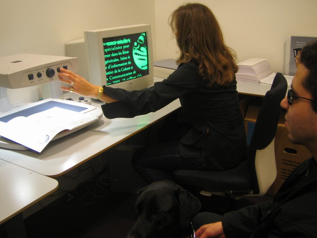 2 personnes dans une ancienne loge de la Bpi. Une bénévole fait une démonstration du télé-agrandisseur à une personne en situation de handicap visuel.