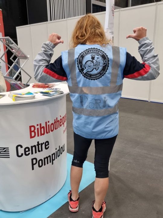 Photo de Dora Balagny de dos qui montre son gilet bleu de Cyclobiblio 2023 devant le stand Bpi du congrès ABF